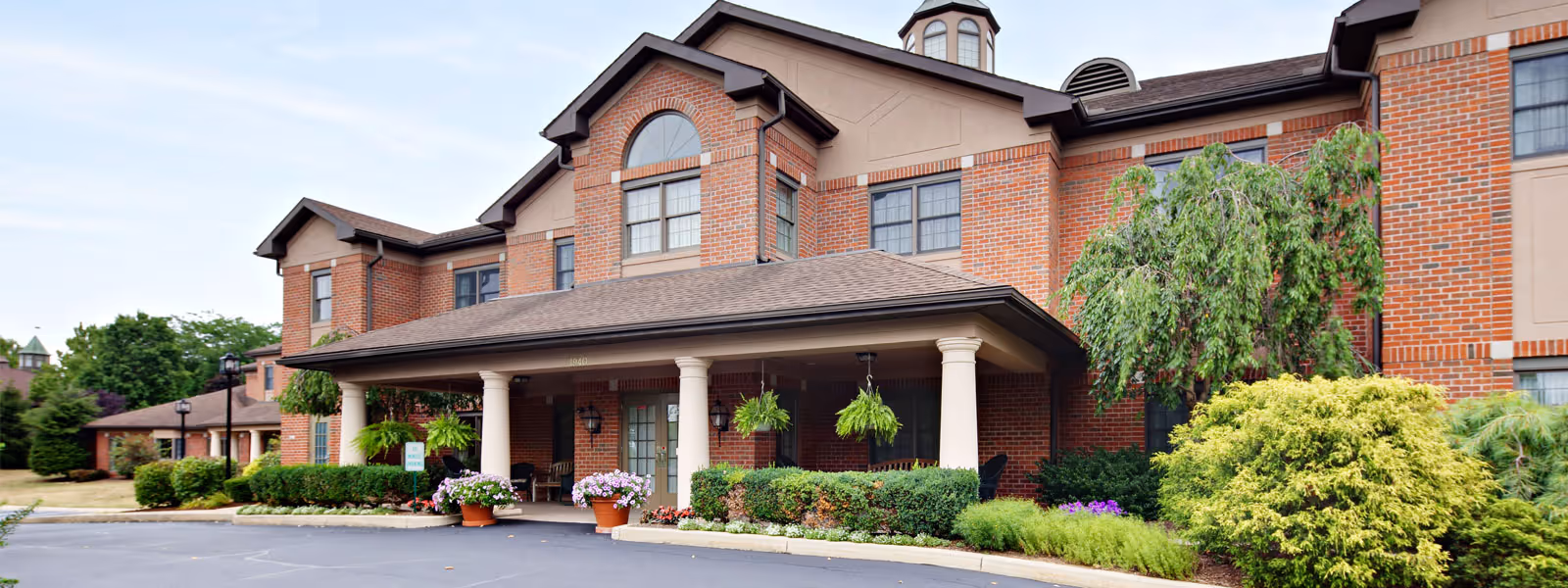 Front exterior view of a senior living facility with a brick facade, large covered entrance supported by white columns, hanging plants, potted flowers, and well-maintained landscaping including bushes and trees.