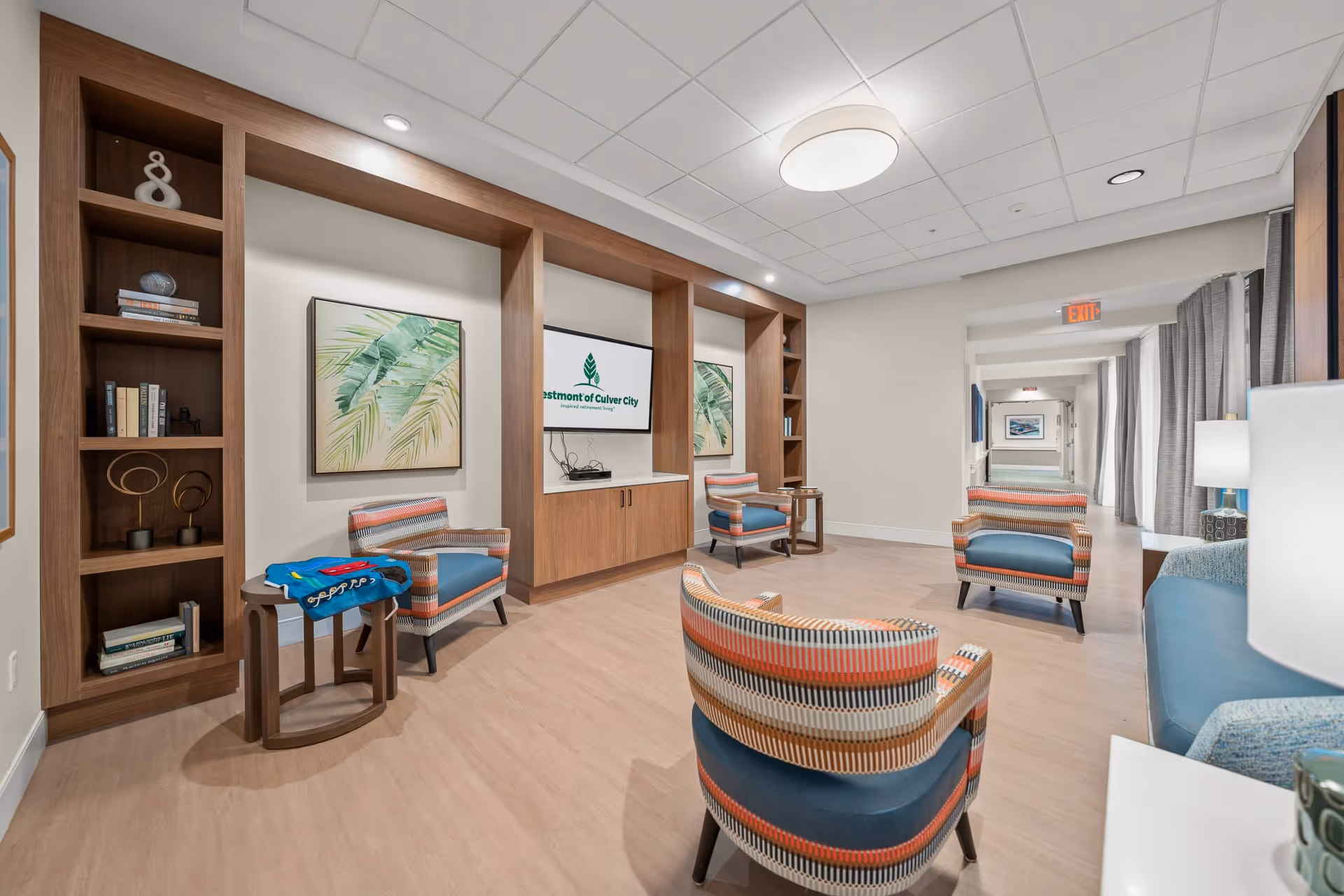 A modern and spacious common area in a senior living facility featuring colorful patterned armchairs, a blue sofa, wooden built-in shelves with decorative items and books, a flat-screen TV displaying the Westmont of Culver City logo, and framed botanical artwork on the walls. The room has light wood flooring, a white ceiling with recessed lighting, and a hallway leading to other parts of the facility.