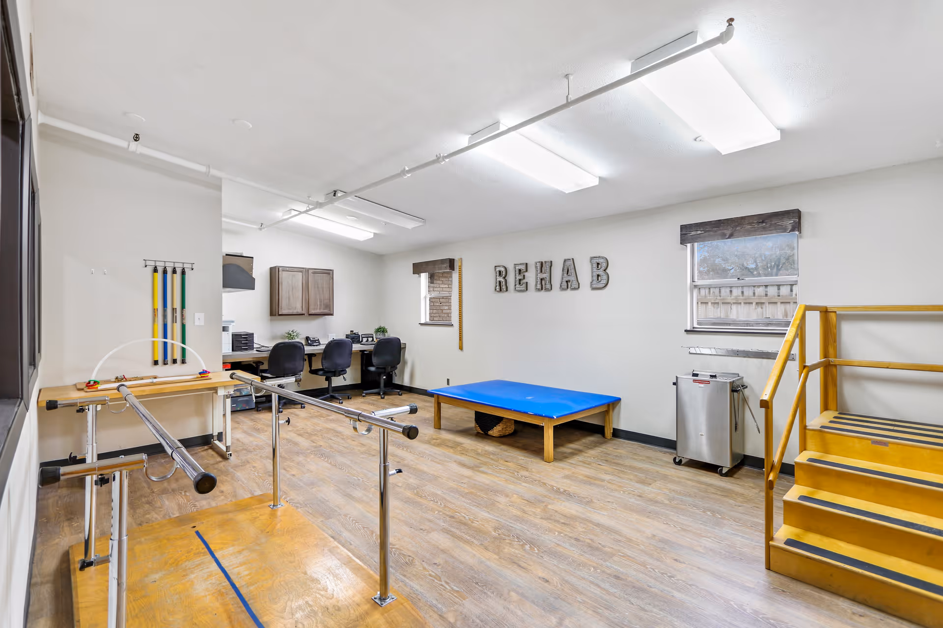 A rehabilitation room with wooden flooring featuring parallel bars for walking exercises, a blue padded therapy table, a set of yellow stairs with handrails, and a desk area with three office chairs and cabinets. The word 'REHAB' is displayed on the wall, and there are two windows with wooden blinds.