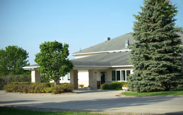 Exterior view of a senior living facility building with a covered entrance, surrounded by trees and shrubs under a clear blue sky.