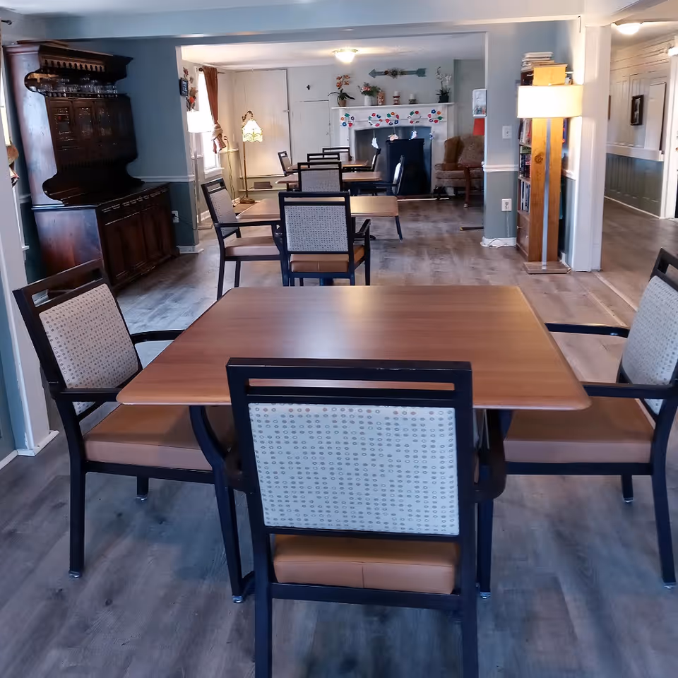 Interior view of a dining area in an assisted living facility with several wooden tables and cushioned chairs arranged neatly. The room has wooden flooring, a vintage wooden cabinet on the left, a fireplace decorated with colorful paper cutouts at the back, and a floor lamp near the right wall. The walls are painted light blue with white trim, and there is a bookshelf with a lamp on the right side.