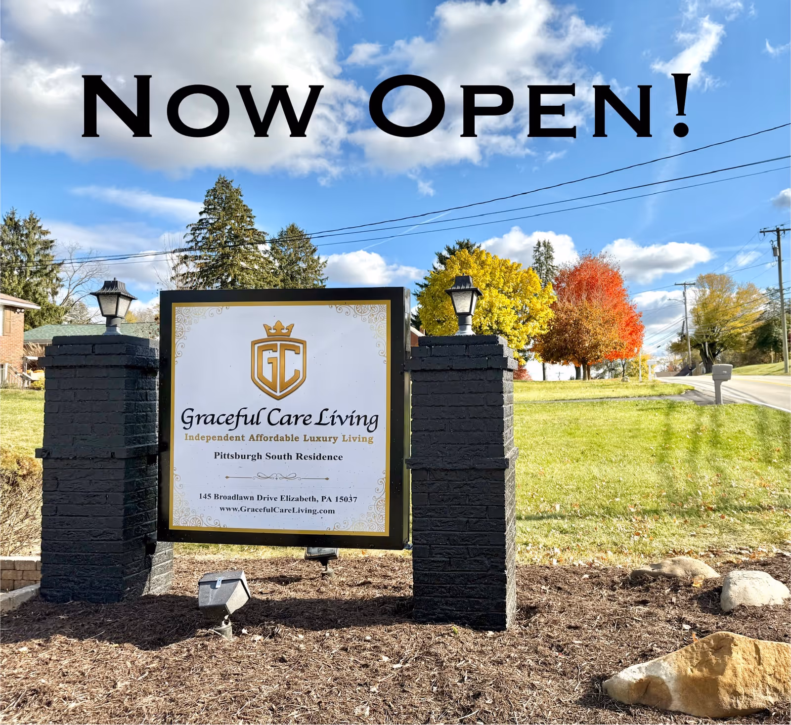Entrance sign reading 'Graceful Care Living' mounted between brick pillars on a grassy lawn with trees and a blue sky.