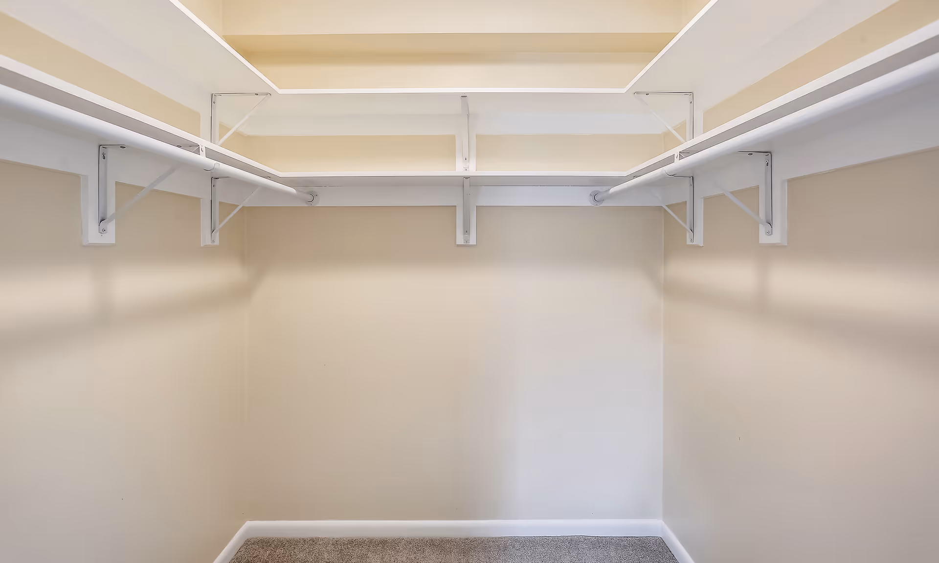 Empty walk-in closet with beige walls, white shelving and hanging rods on both sides, and carpeted floor.