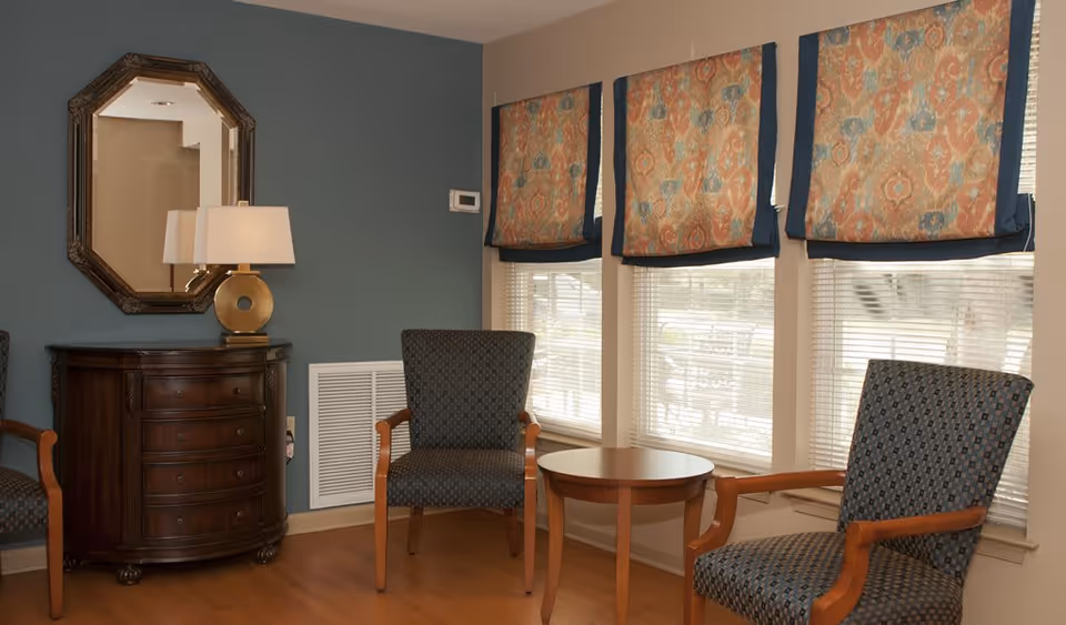 A cozy sitting area with upholstered chairs, a round wooden table, a lamp on a dark wooden dresser, and patterned window shades.