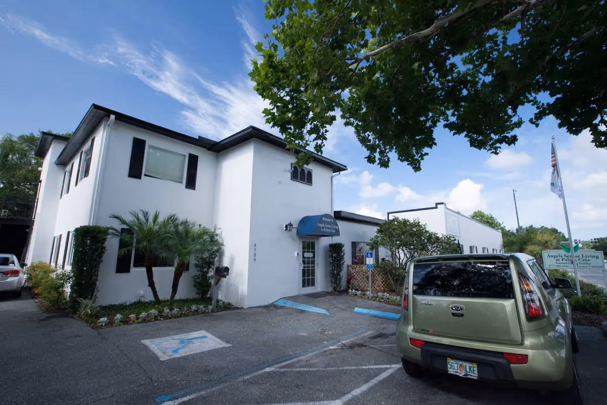 Front exterior of a white two-story senior living building with a small parking lot, handicap spaces, and a green car parked near the entrance.