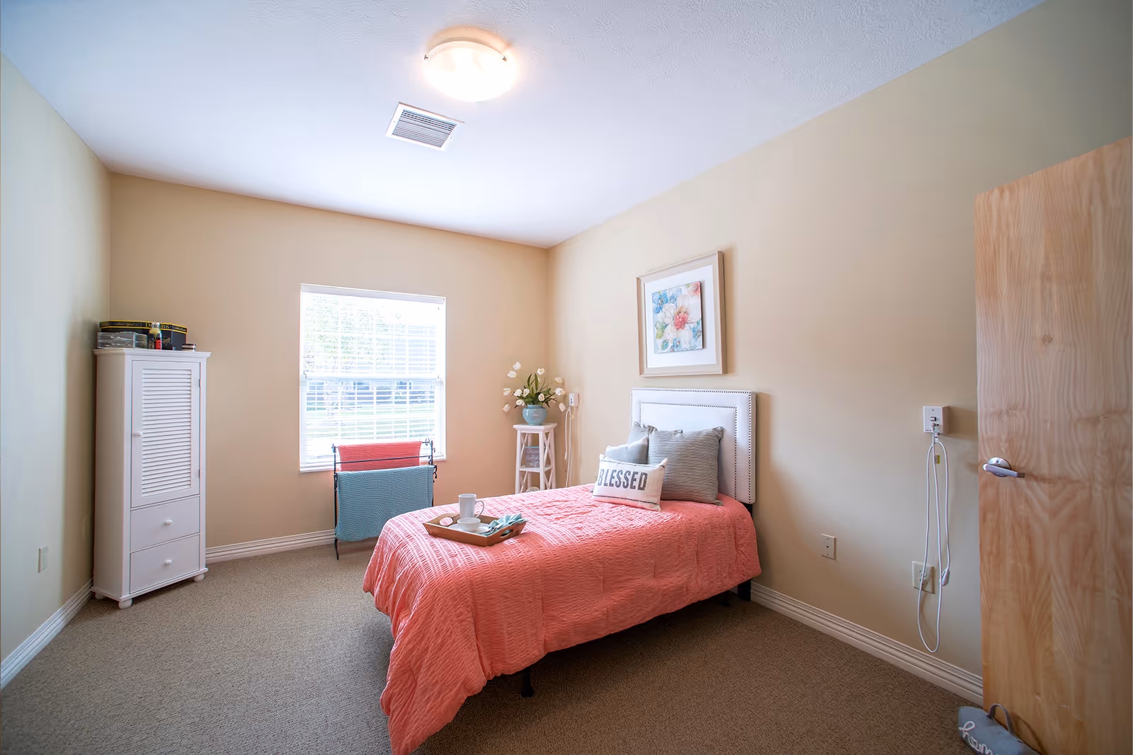 A cozy bedroom with beige walls and carpeted floor featuring a single bed with a coral pink quilt and decorative pillows, including one that says 'BLESSED'. A tray with a cup and napkin is placed on the bed. There is a window with white blinds letting in natural light, a white cabinet in the corner, a small stand with a vase of flowers, and a wooden door slightly ajar.