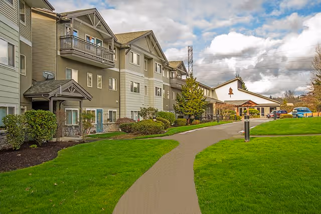 Exterior view of a multi-story senior living facility with beige and green siding, balconies, and a well-maintained lawn with a paved walkway leading to the entrance. The sky is partly cloudy.