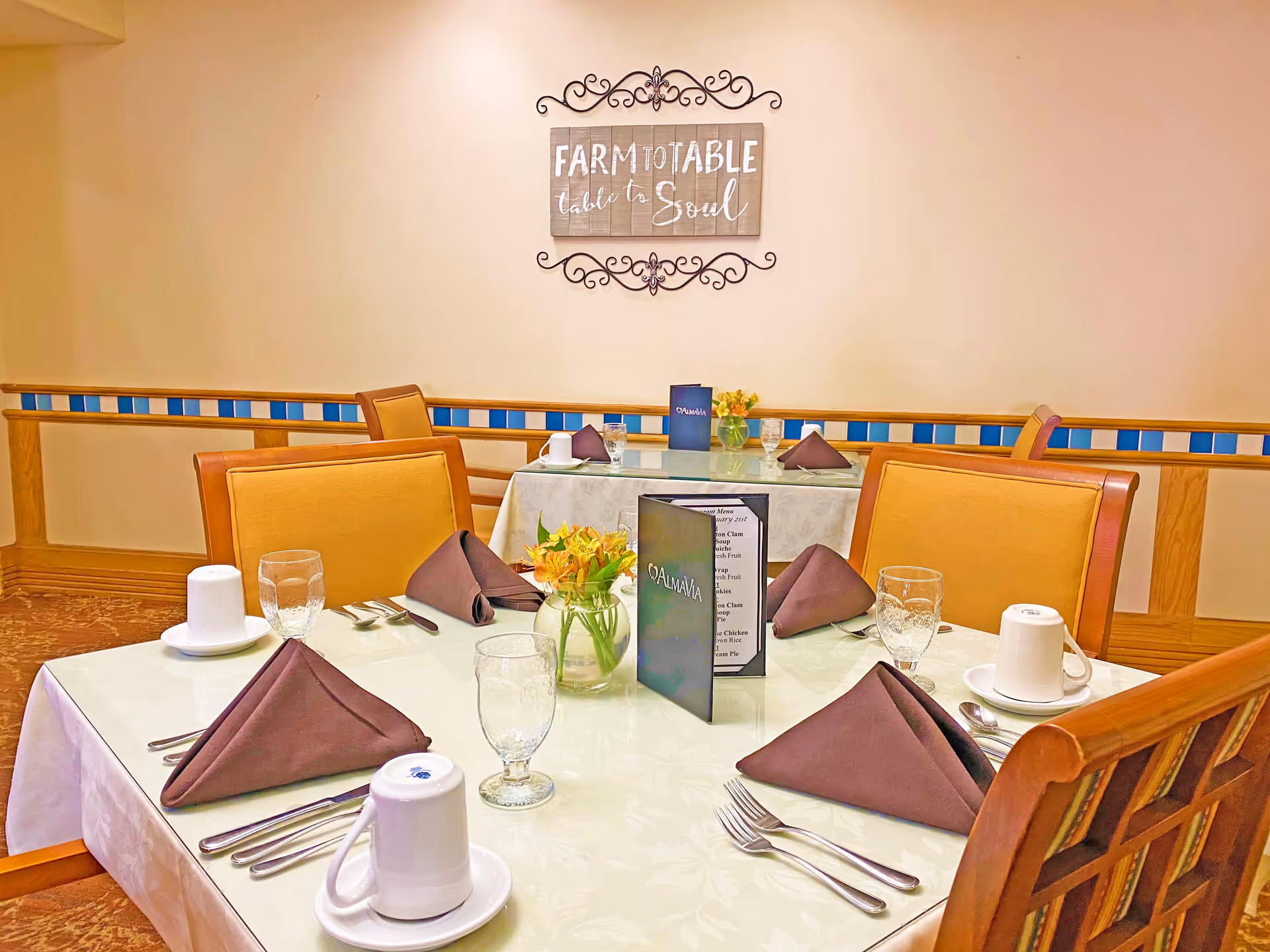 A dining room table set for four with brown folded napkins, white cups on saucers, water glasses, silverware, and a small vase with yellow flowers. Another similarly set table is visible in the background. On the wall above the tables is a decorative sign that reads 'FARM TO TABLE table to Soul'.