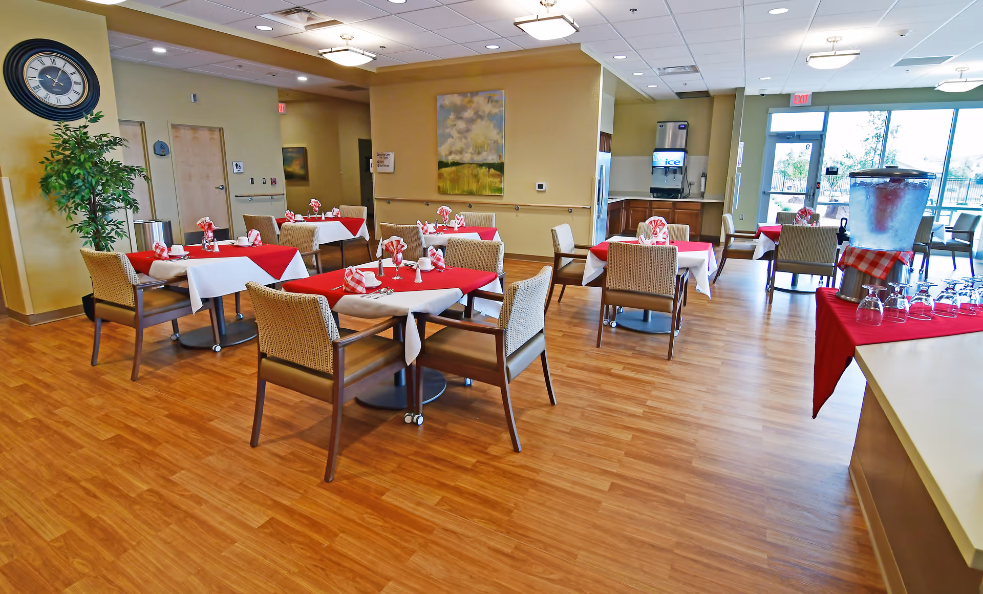 A dining room with several tables covered with red and white tablecloths, each set with napkins and utensils. The room has wooden flooring, beige walls, a large clock on the wall, a potted plant, and a beverage station with glasses and a large water dispenser. Large windows and a glass door allow natural light to enter the space.