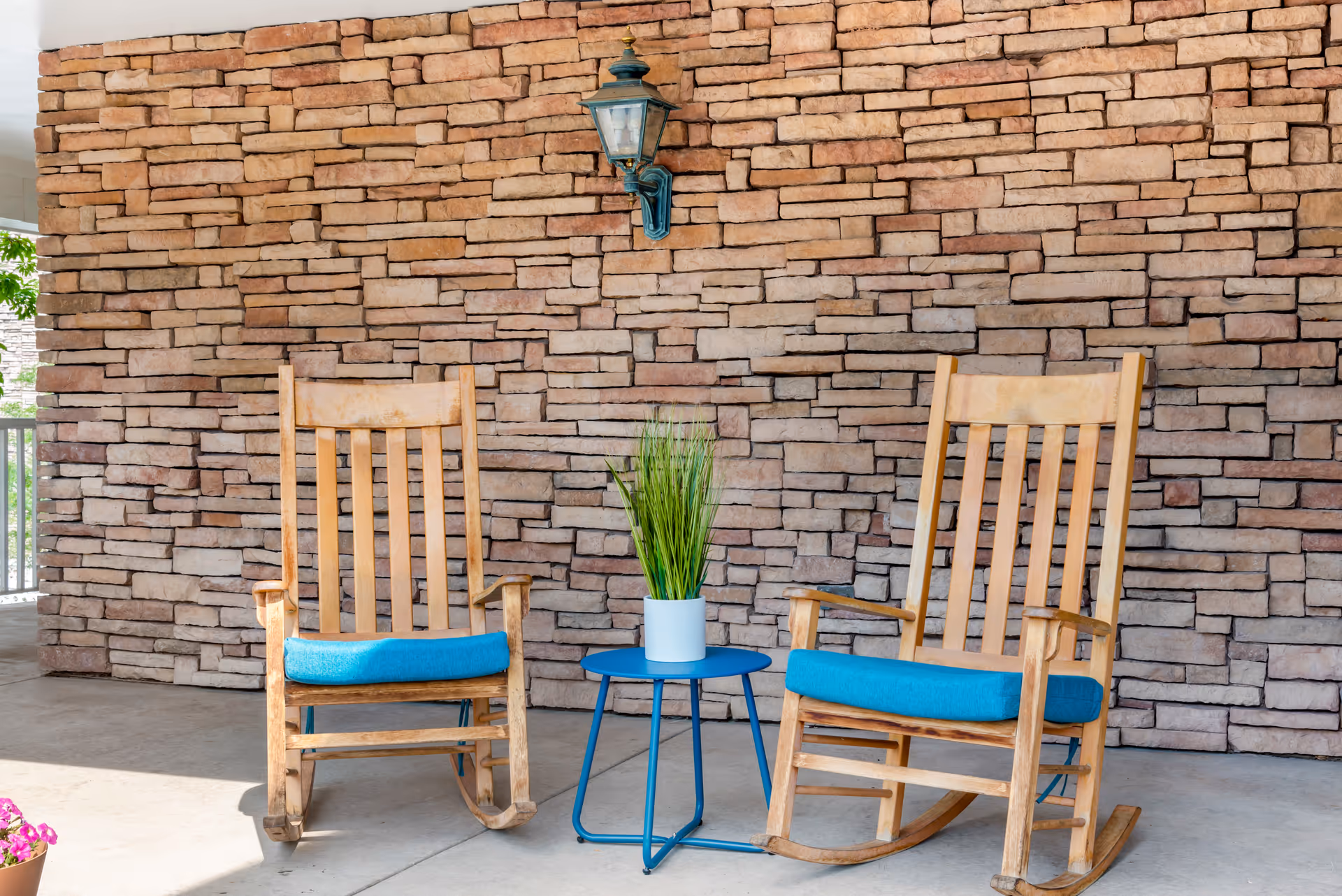 Two wooden rocking chairs with blue cushions are placed on a concrete floor in front of a stone brick wall. Between the chairs is a small round black metal table with a white pot containing green grass-like plants. A vintage-style wall lantern is mounted on the stone wall above the table.