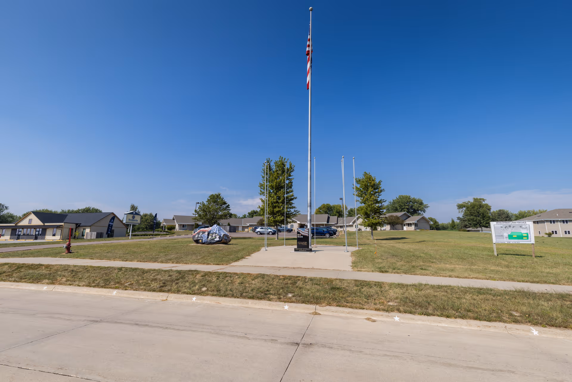 Wide exterior view of Holstein Senior Living facility showing a flagpole with an American flag in the center of a grassy area, surrounded by buildings, trees, a painted rock, and a sign with facility information under a clear blue sky.