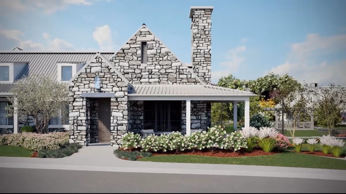 Stone building with a chimney and a covered porch surrounded by well-maintained landscaping including bushes, flowers, and trees under a partly cloudy sky.