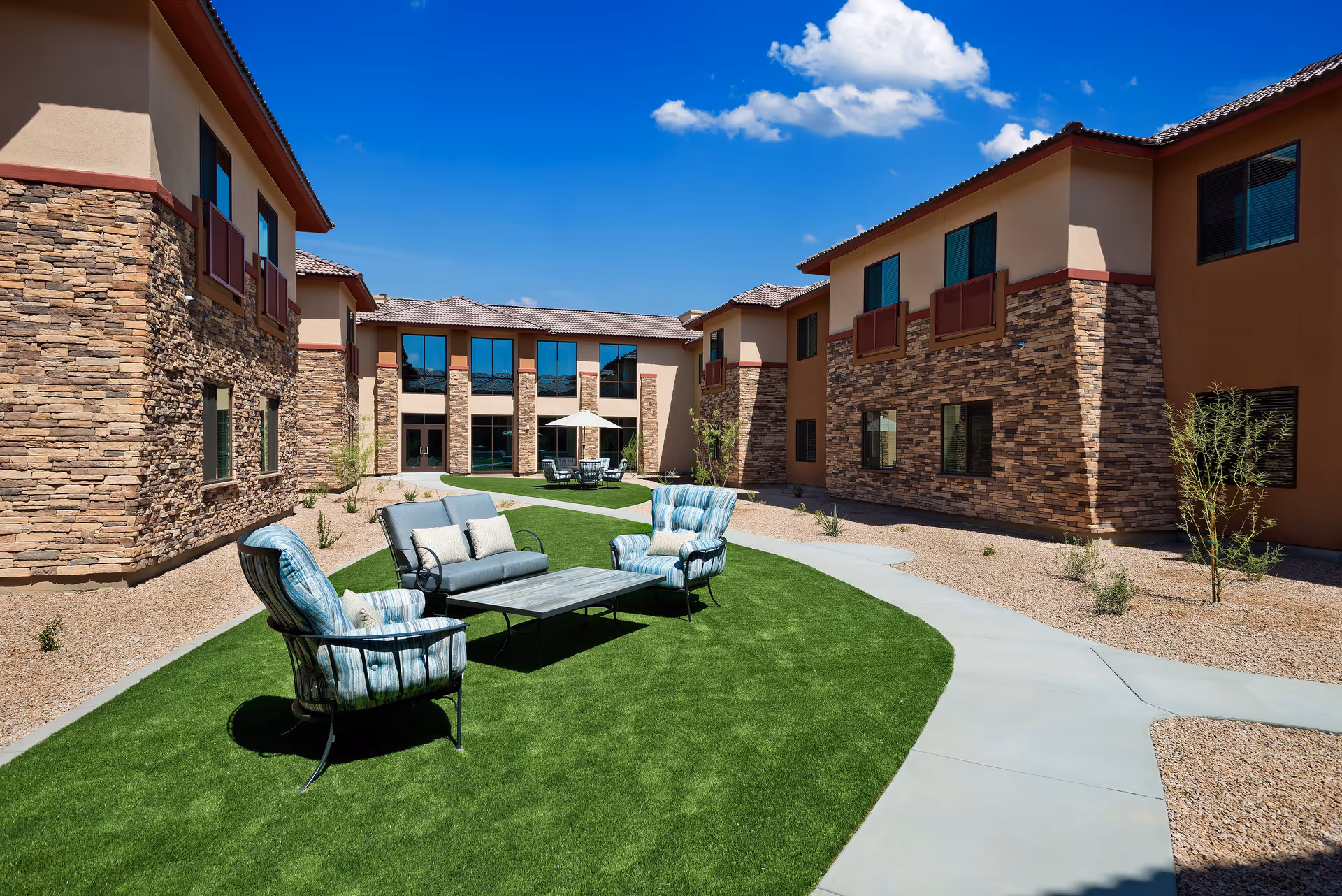 Outdoor courtyard area of a senior living facility with stone and stucco buildings surrounding a green artificial lawn. There are cushioned patio chairs and a loveseat arranged around a coffee table on the lawn, with a concrete walkway curving through the space. The sky is clear and blue with a few clouds.