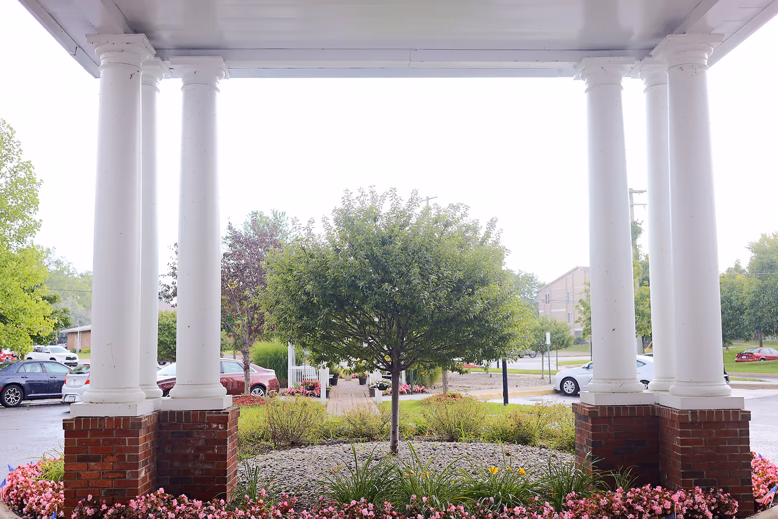 View from under a covered porch with white columns and brick bases, looking out onto a landscaped area with a small tree, bushes, and flowers. In the background, there is a parking lot with several cars and more greenery.