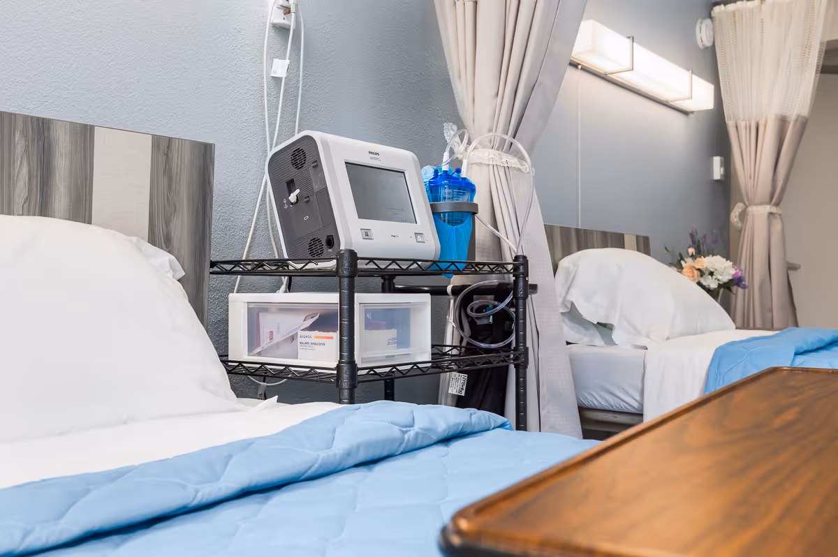 Two hospital-style beds with light blue blankets and white pillows in a care facility room. Between the beds is a black metal cart holding medical equipment and supplies. The room has light gray walls, a large mirror, and curtains for privacy. A wooden overbed table is visible in the foreground.