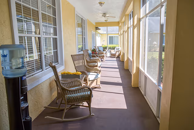A sunlit screened-in porch with several wicker rocking chairs and small tables, some with flower arrangements. There is a water dispenser on the left side near the windows, and ceiling fans are mounted overhead. The porch overlooks a garden area outside the screened windows.