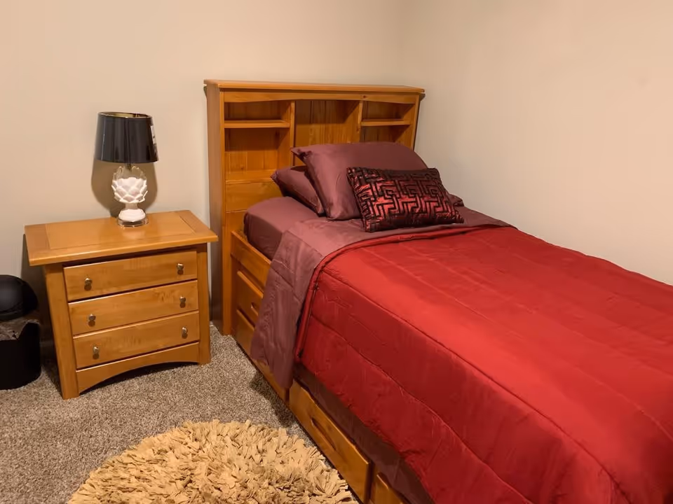 A small bedroom with a single wooden bed featuring built-in shelves in the headboard. The bed is made with red and maroon bedding and pillows. Next to the bed is a wooden nightstand with three drawers and a white lamp with a black lampshade. The floor is carpeted and there is a round beige rug partially visible.