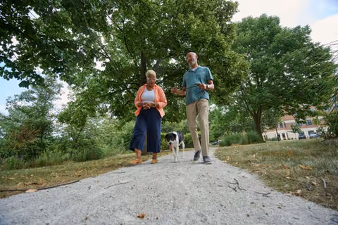 Two people walking a dog along a tree-lined path in a grassy outdoor area.