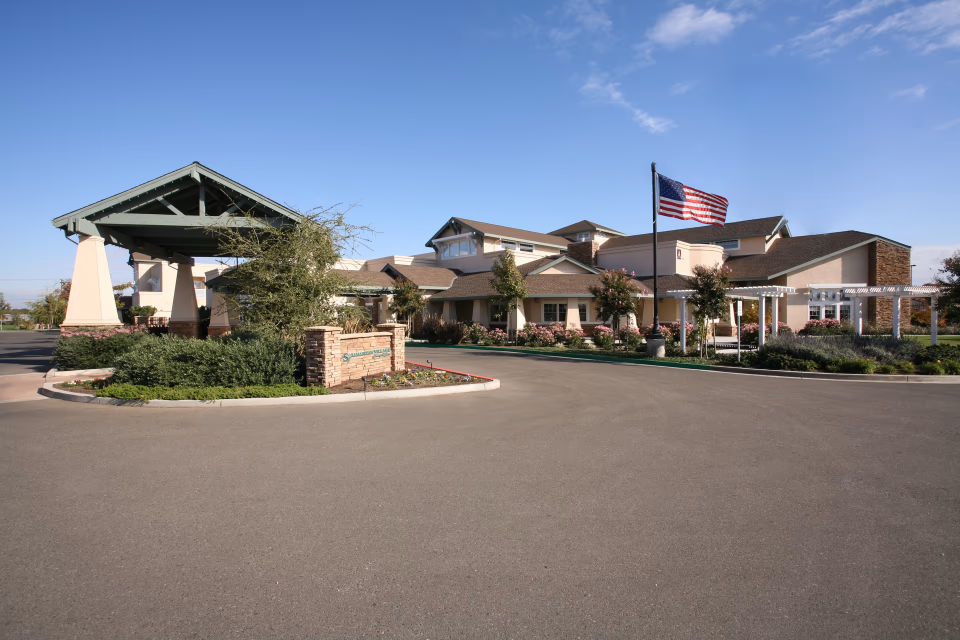 Exterior view of Samaritan Village facility showing a large building with a covered entrance, landscaped greenery, an American flag on a flagpole, and a clear blue sky.