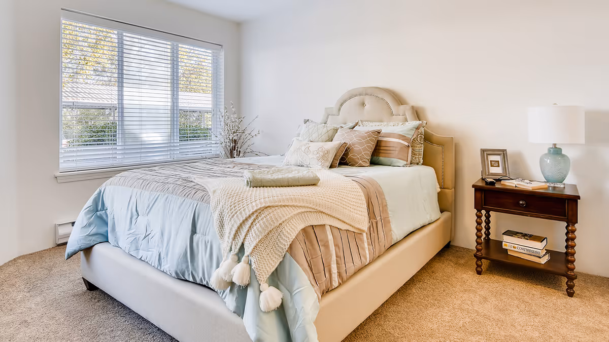 A neatly made bed with multiple pillows and a knitted throw blanket in a bright bedroom with a large window and a wooden nightstand holding a lamp, picture frame, and books.
