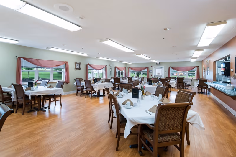 A spacious dining room with multiple tables covered in white tablecloths, each set with napkins, cups, glasses, and small flower vases. The room has large windows with red curtains, wooden chairs, and a light wood floor. There is a beverage station on the right side of the room.