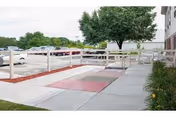 Outdoor view of a paved walkway with handrails leading to the entrance of a building, surrounded by landscaped areas with mulch and plants, and a parking lot with several cars in the background under a cloudy sky.
