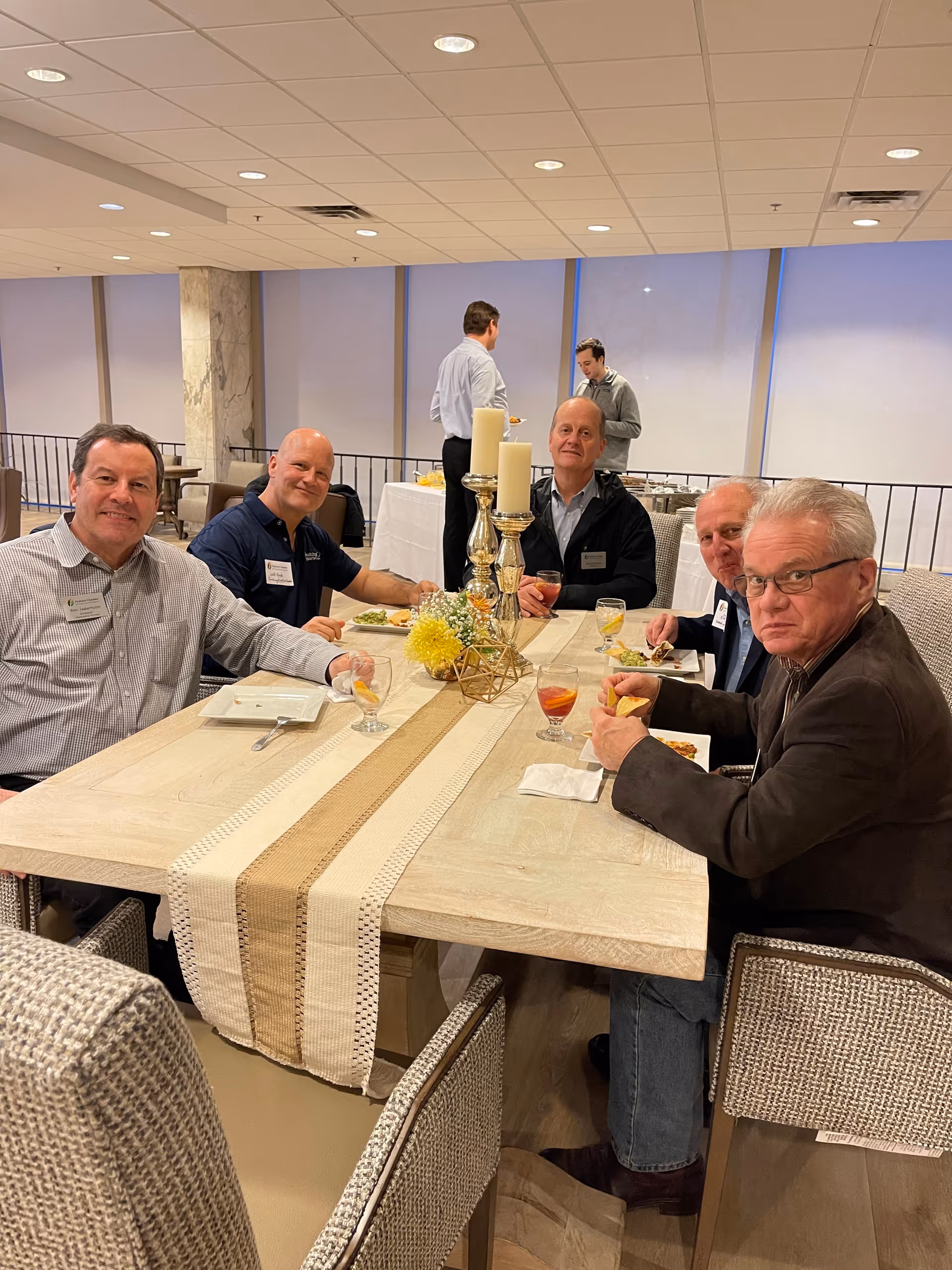 Five men sitting around a rectangular dining table with a beige table runner, candles, and a flower centerpiece, eating and drinking in a well-lit indoor setting. Two other men are standing in the background near a buffet table.
