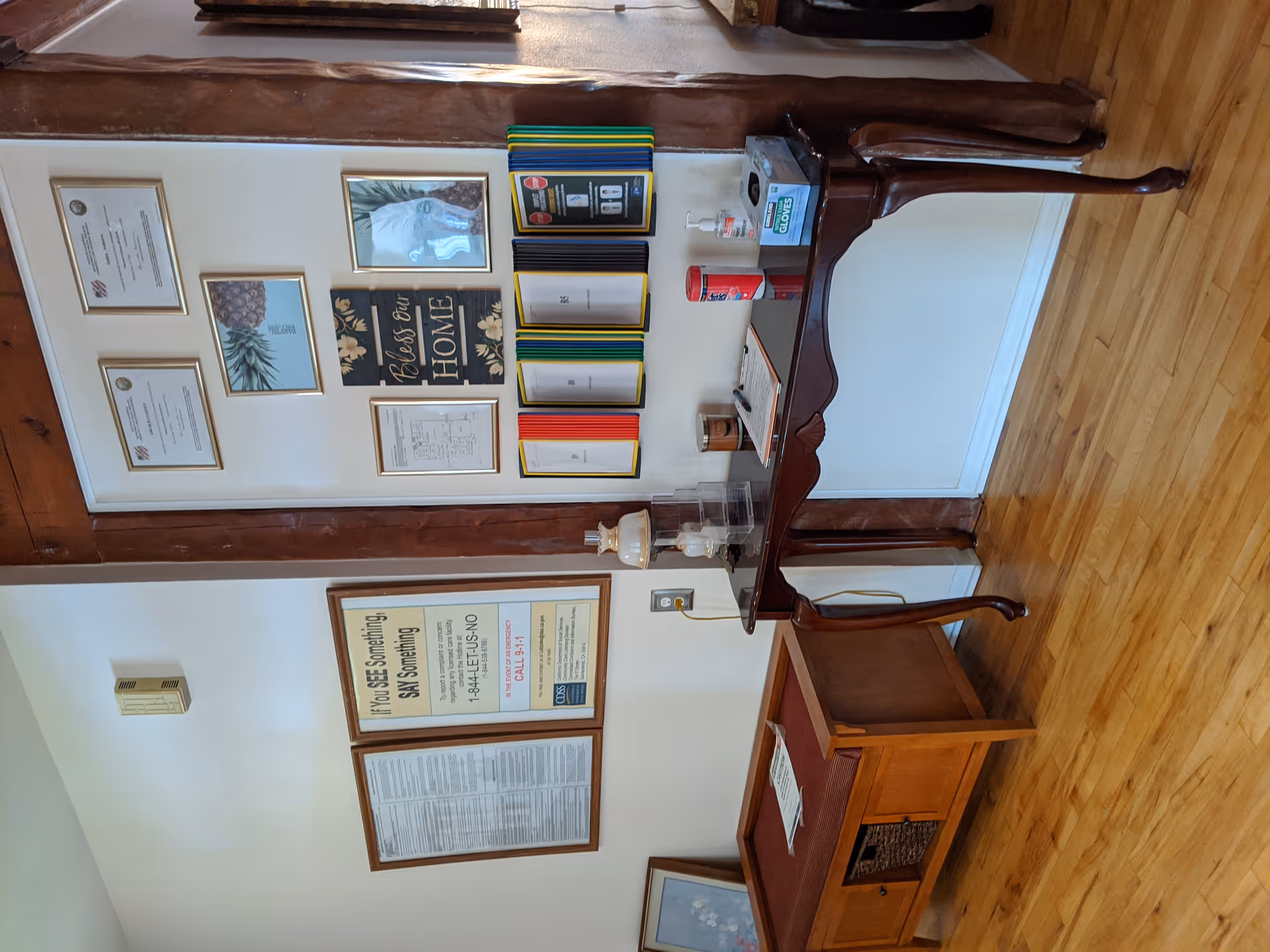 Interior view of a senior living facility corner with a wooden table holding a clipboard, hand sanitizer, disinfecting wipes, and a box of gloves. Above the table are framed certificates, a pineapple picture, a 'Bless Our Home' sign, and several colorful folders mounted on the wall. To the left, there is a wooden bench with a cushion and framed notices on the wall.