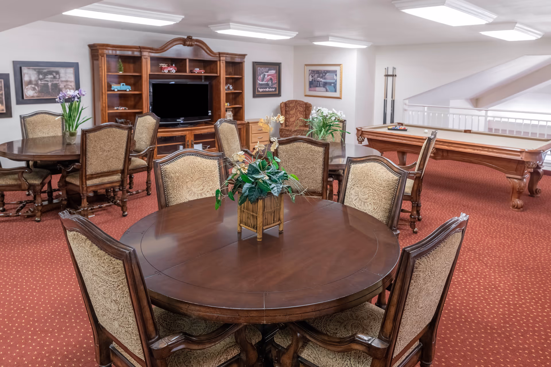 A cozy common area with round wooden tables and upholstered chairs arranged around them. There is a wooden entertainment center with a TV and decorative items, framed pictures on the walls, a pool table with billiard balls, and a red carpeted floor. The room is well-lit with ceiling lights.