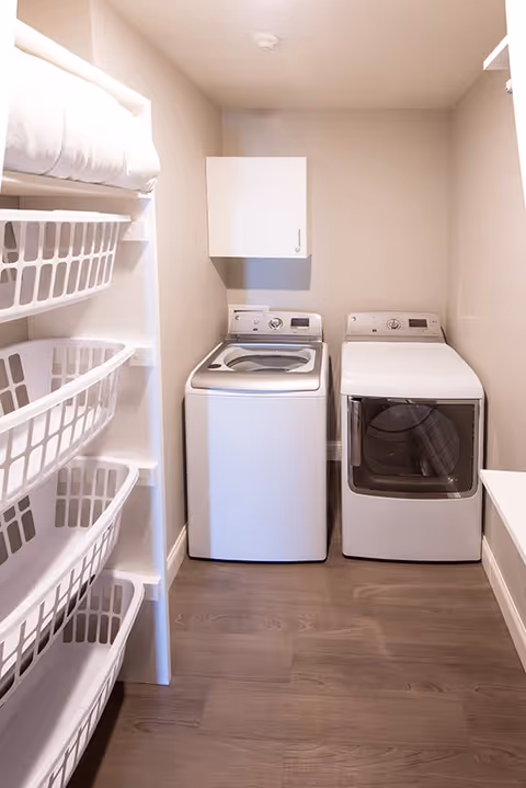 A laundry room with a washing machine and a dryer side by side against a beige wall. To the left, there are white shelves holding multiple white laundry baskets. The floor is wooden, and there is a small white cabinet mounted on the wall above the washing machine.