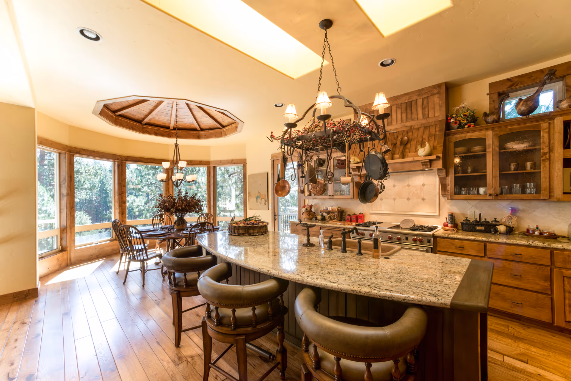 A spacious kitchen and dining area with large windows offering a view of trees outside. The kitchen features a large granite island with three cushioned bar stools, a hanging pot rack with various pots and pans, wooden cabinets, and a stove with a wooden hood. The dining area has a round wooden table with four chairs and a chandelier above it. The floor is wooden, and the ceiling has a recessed wooden design above the dining table.