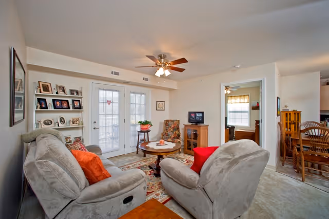 A cozy living room with two beige recliner chairs featuring orange and red pillows, a round wooden coffee table with a plant and decorative items, a floral armchair, a small TV on a wooden stand, and a white shelf with framed photos. French doors with glass panes lead outside, and a ceiling fan with lights is mounted on the ceiling. The room opens into a dining area with a wooden dining table and chairs.