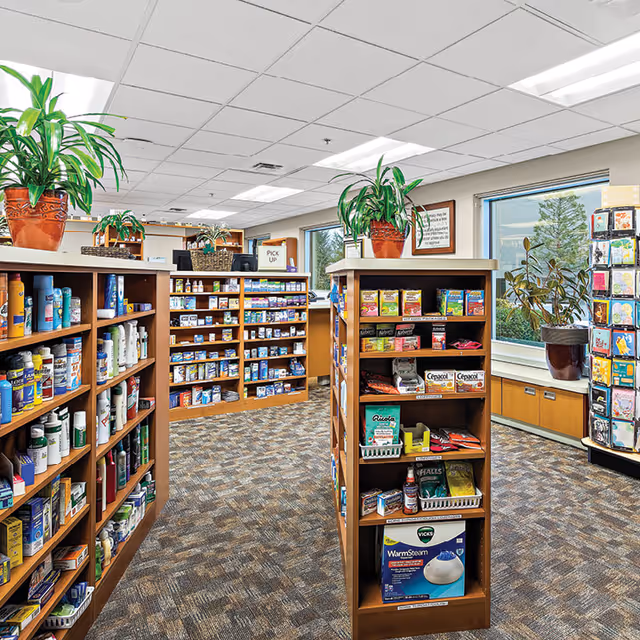 Interior view of a well-lit retail or convenience area with wooden shelves stocked with various health and wellness products, including lotions, medicines, and personal care items. There are large windows with potted plants on the windowsills, and the floor is carpeted with a patterned design.