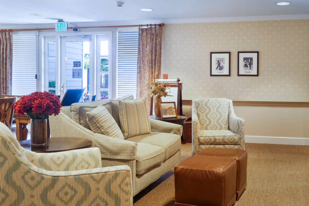 A cozy senior living facility lounge area with a beige sofa and two patterned armchairs. There are two brown leather ottomans in front of the seating. A round wooden side table with a vase of red flowers is next to one armchair. The room has beige wallpaper with a subtle pattern, two framed black and white photos on the wall, and large windows with curtains letting in natural light. An exit door is visible in the background.