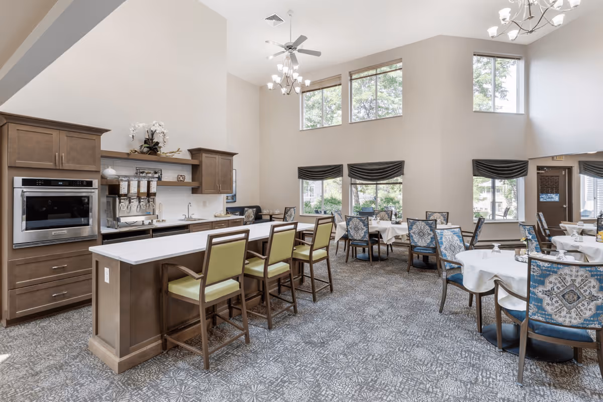 A bright and spacious dining area in a senior living facility featuring multiple round tables with white tablecloths and blue patterned chairs. There is a kitchen counter with green cushioned bar stools, wooden cabinetry, a built-in oven, and a beverage dispenser. Large windows with dark valances allow natural light to fill the room, and chandeliers hang from the high ceiling.