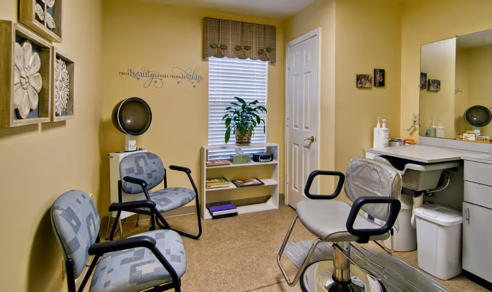 A small salon or beauty room with three chairs, including a salon chair with armrests and a hair dryer hood. The walls are painted yellow and decorated with floral wall art and a quote that reads 'True beauty comes from within.' There is a window with blinds and a valance, a small white bookshelf with magazines and a plant, and a countertop with a sink, soap dispensers, and a mirror.