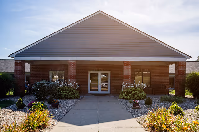 Covered entrance of a brick senior living facility with double glass doors and a landscaped walkway.