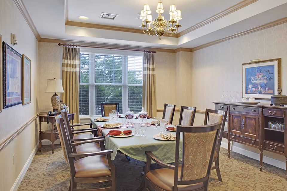 A formal dining room with a long table covered with a green tablecloth, set with plates, glasses, and napkins. The room has eight wooden chairs with upholstered seats and backs. There is a large window with curtains letting in natural light, a chandelier hanging from the ceiling, a sideboard with glassware and a framed painting on the wall, and a table lamp on a small side table in the corner.