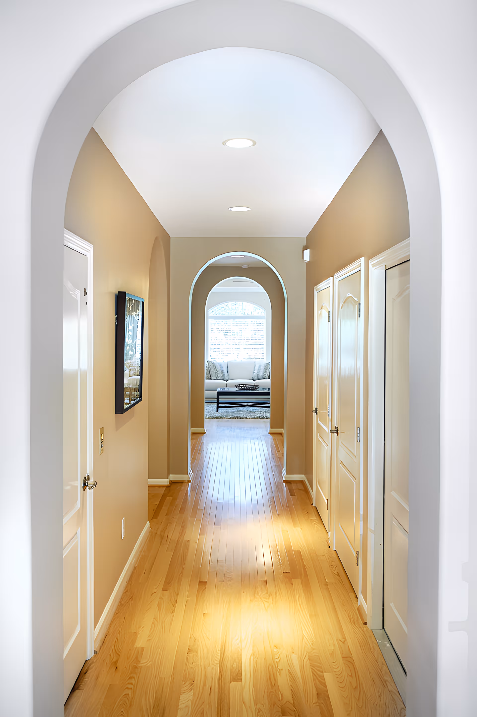 A bright hallway with light wooden flooring and beige walls, featuring multiple white doors on the right side and an arched doorway at the end leading to a living room with a white sofa and a coffee table in front of a large window.