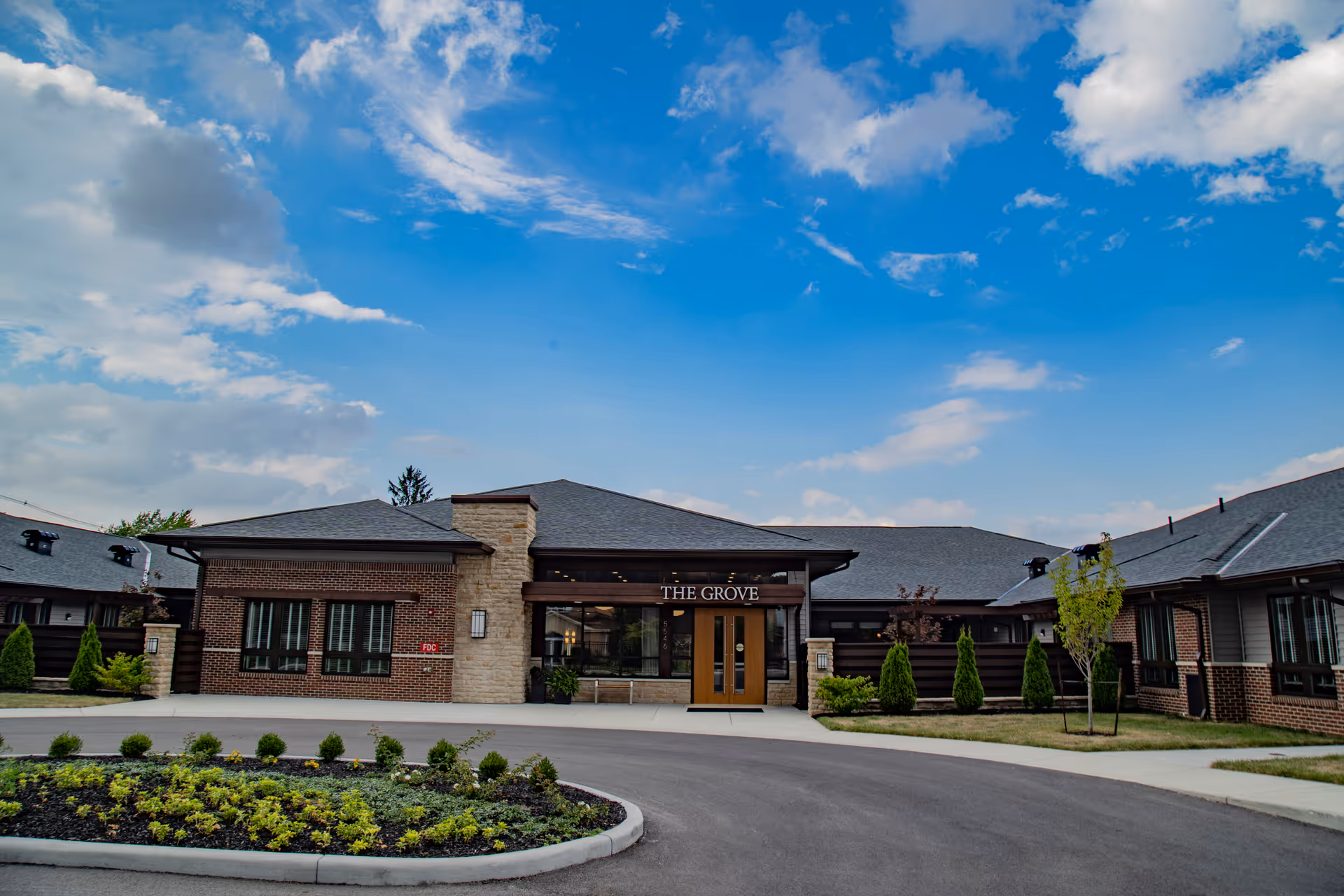 Exterior view of The Grove at Oakleaf Village of Columbus, showing a single-story brick and stone building with a central entrance under a sign that reads 'THE GROVE'. The building is surrounded by landscaped greenery and a circular driveway, under a partly cloudy blue sky.