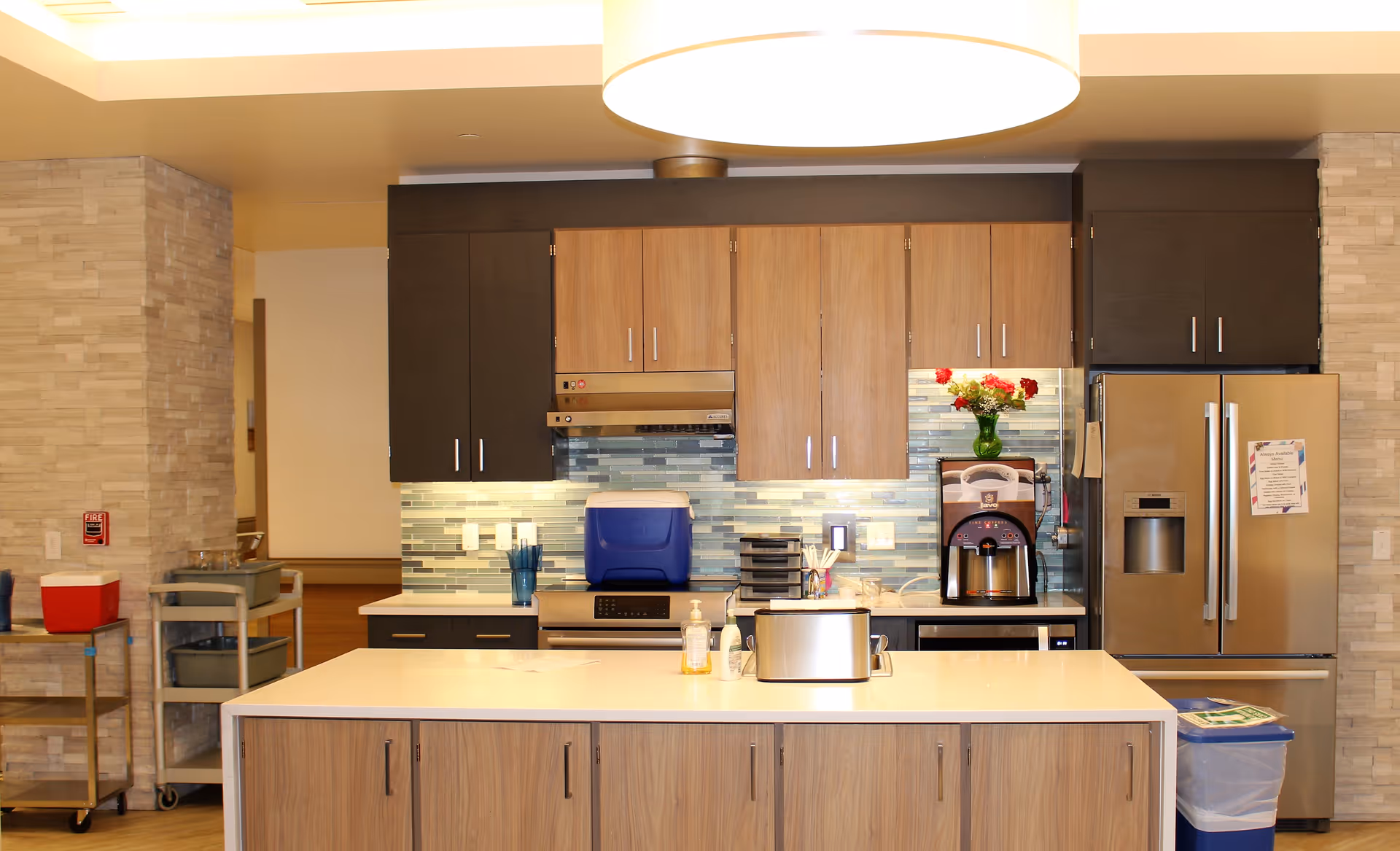 Modern communal kitchen with a central island, wooden cabinets, stainless steel refrigerator, and coffee machine.