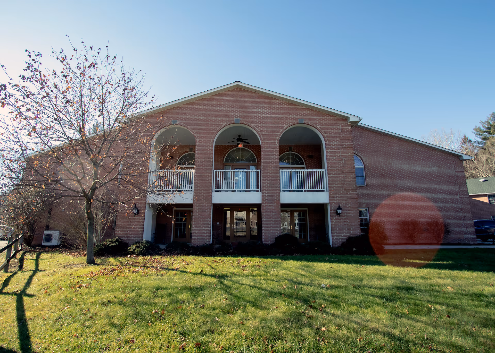 Front exterior view of a two-story brick building with three large arched openings on the upper level, white railings, and a green lawn with a leafless tree in front under a clear blue sky.