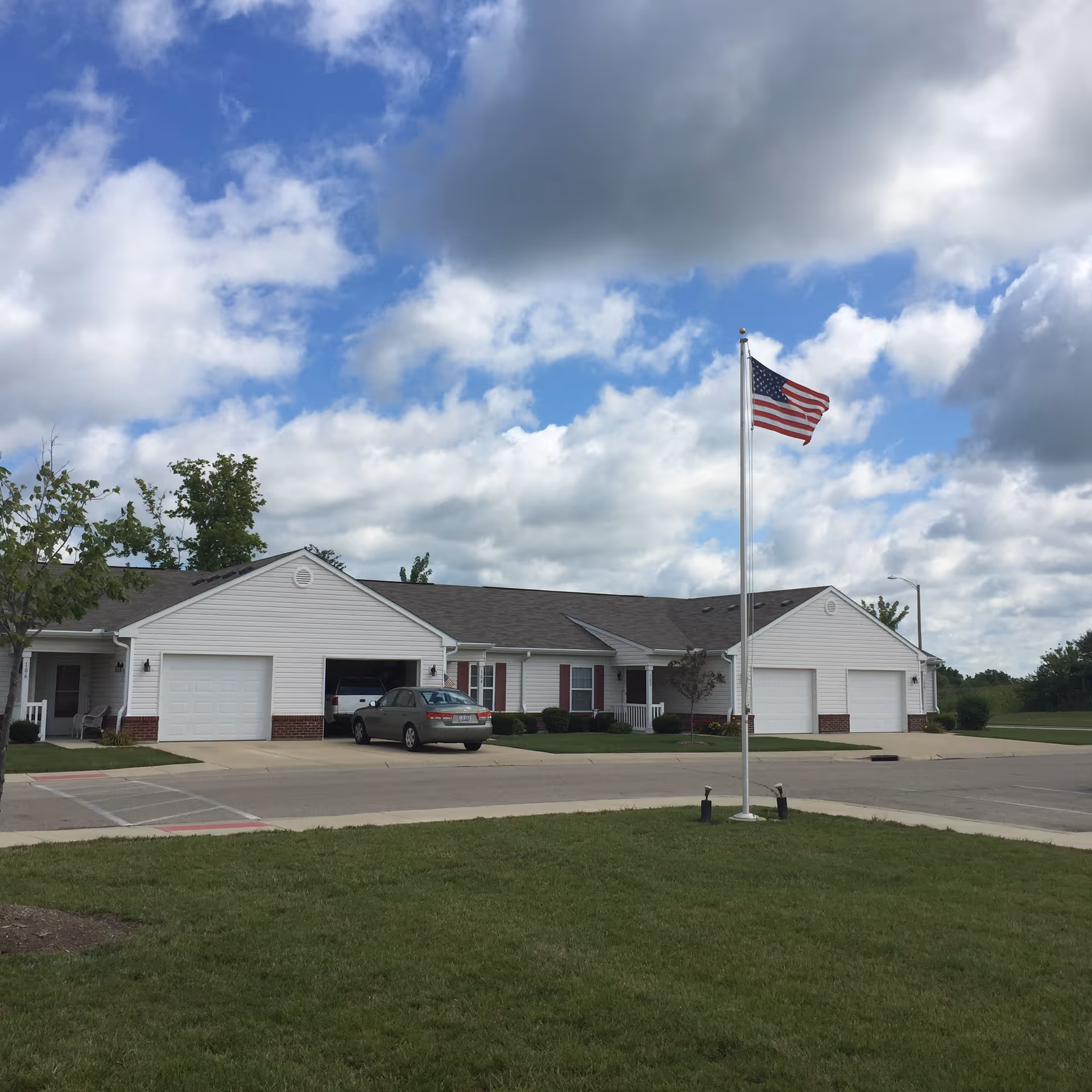 Single-story white senior living building with attached garages and an American flag on a pole under a partly cloudy sky.