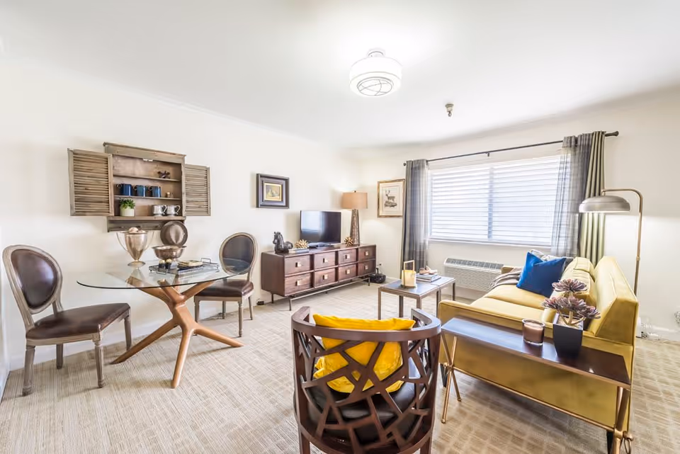 A bright living room with a yellow sofa, a wooden chair with a yellow cushion, a glass-top round dining table with two brown chairs, a wooden TV stand with a flat-screen TV, a floor lamp, and a window with blinds and curtains.