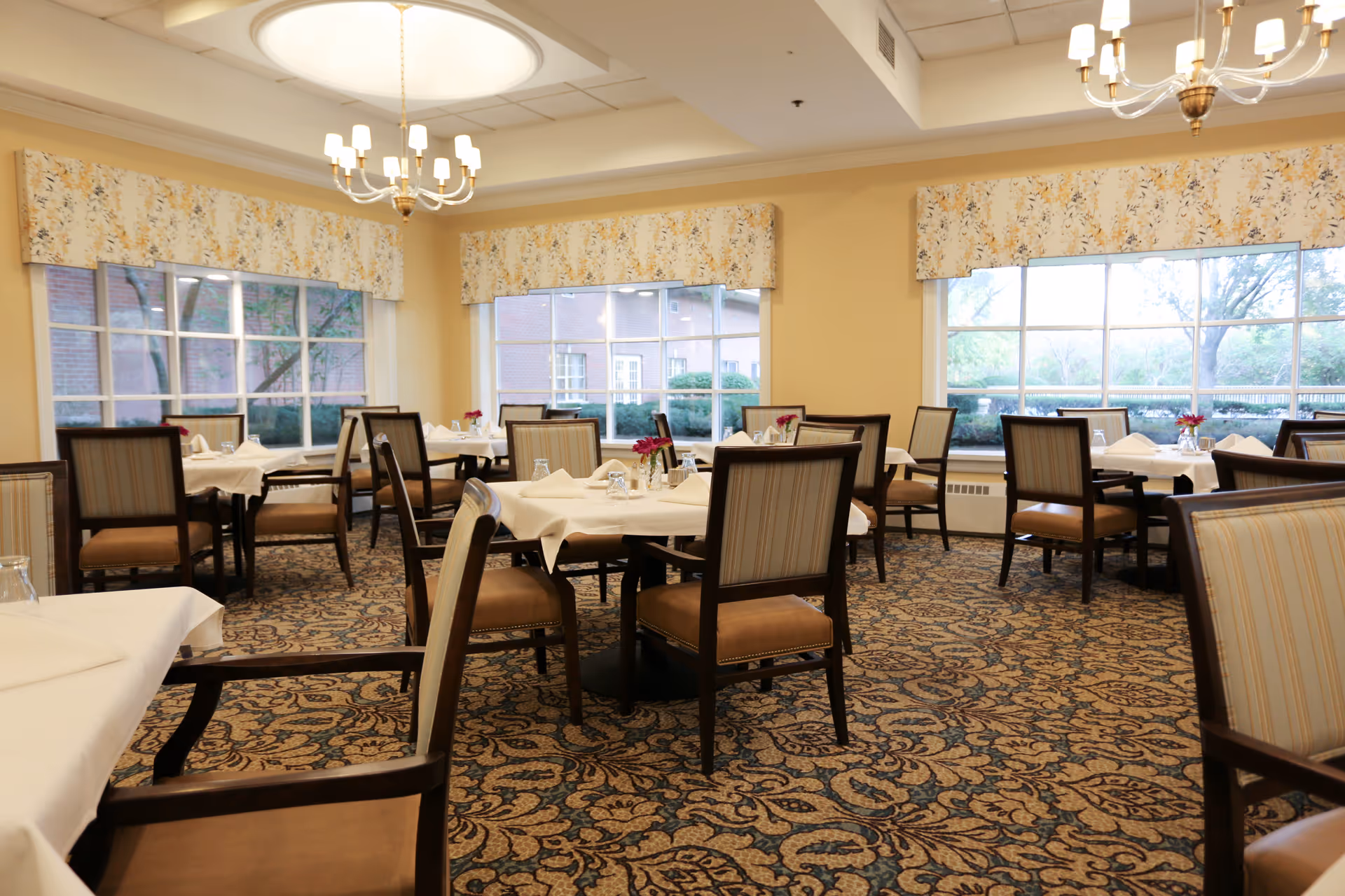 A well-lit dining room with multiple tables covered in white tablecloths, each set with glasses and napkins. The room features large windows with floral valances, patterned carpet, and wooden chairs with cushioned seats and backs. Two chandeliers hang from the ceiling providing warm lighting.