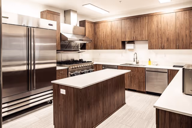 Modern kitchen with stainless steel appliances including a large refrigerator, stove with range hood, dishwasher, and microwave. The kitchen features wooden cabinets, a central island with a white countertop, and a sink with a faucet. The floor is tiled and the room is well-lit with ceiling lights.