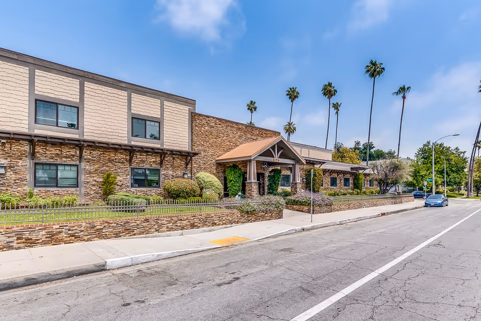 Exterior view of a senior living facility building with stone and beige siding, a covered entrance with the number 925, landscaped bushes and flowers, palm trees in the background, and a street with parked cars in front.