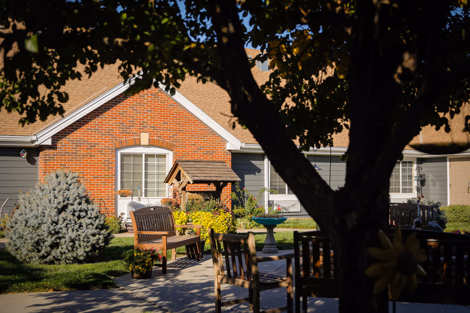 Shaded courtyard with wooden benches, a birdbath and landscaping in front of a brick-faced assisted living building.