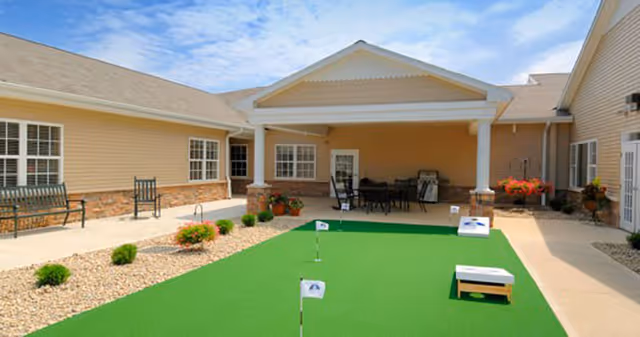 Outdoor courtyard area at Westlake Health Campus featuring a putting green with small flags, surrounded by beige buildings with windows and a covered patio area with tables and chairs. There are benches and potted plants along the pathways.