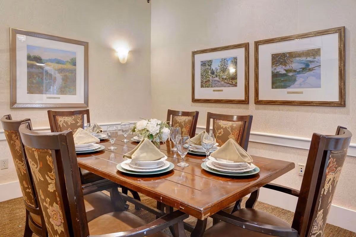 A dining room with a wooden table set for six people. Each place setting includes a plate, bowl, folded beige napkin, and a water glass. The chairs have dark wooden frames with floral upholstered backs. Three framed landscape paintings hang on the beige walls, and a small floral centerpiece is on the table.